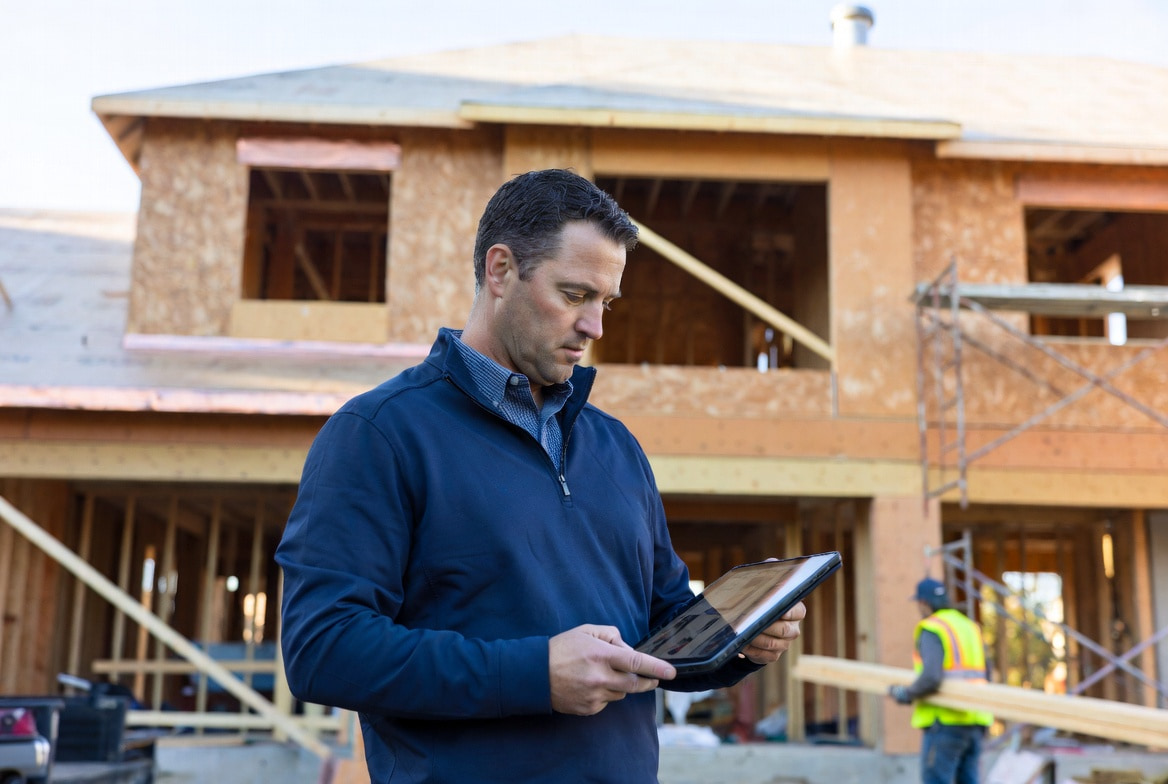 General contractor reviewing AI project management software on a tablet at a residential construction site with subcontractors working in the background, illustrating the 2026 AI software stack for general contractors across small, mid, and large GC operations