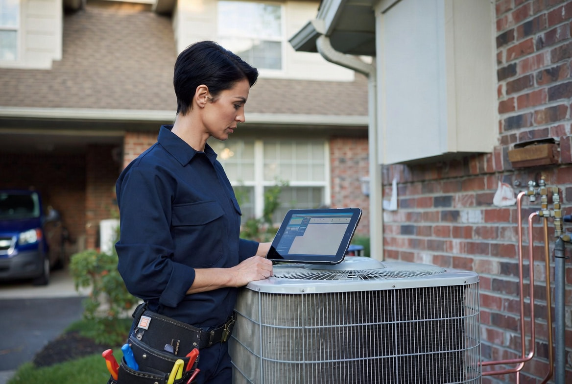 HVAC technician reviewing AI field service management software on a tablet next to a residential condenser unit, illustrating the 2026 AI tool stack for HVAC contractors